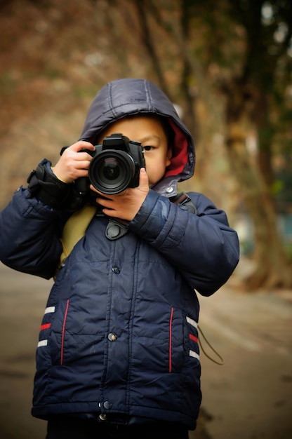 Foto jongen in warme kleding die met een camera fotografeert terwijl hij tegen bomen staat