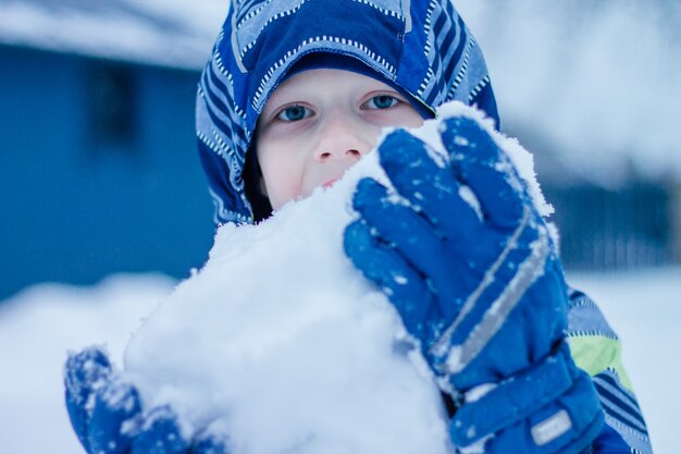 Jongen in winteroverall staat buiten en eet sneeuw, met een grote klomp sneeuw in zijn handen