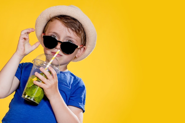 Jongen in zomeroutfit die een zonnebril draagt en geniet terwijl hij een Mojito-cocktail drinkt. Poseren op de gele achtergrond.