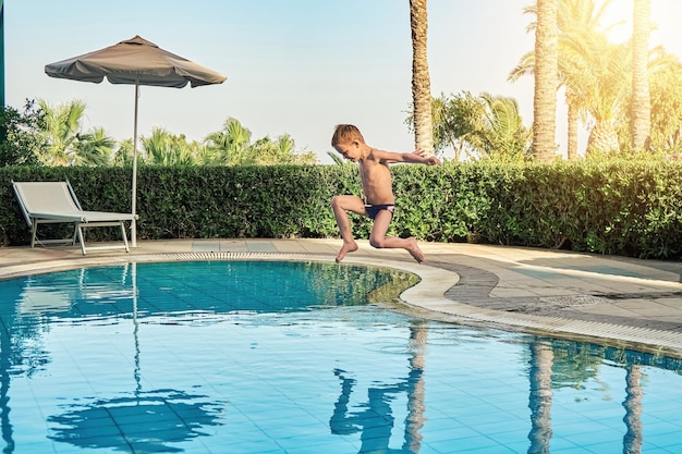 Jongen in zwembroek springen in kinderbad tegen ligbedden en palmbomen in luxehotel