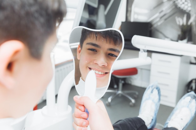 Jongen kijkt in de spiegel met een brede glimlach zittend op een stoel met een tandarts in de tandartspraktijk