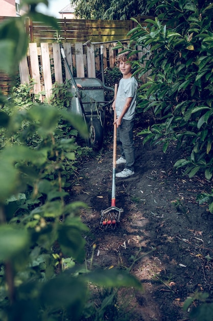 Jongen kijkt naar camera terwijl hij in de groene tuin staat in de buurt van houten hek en aangrijpende grond met tuincultivator in zonnige zomerdag