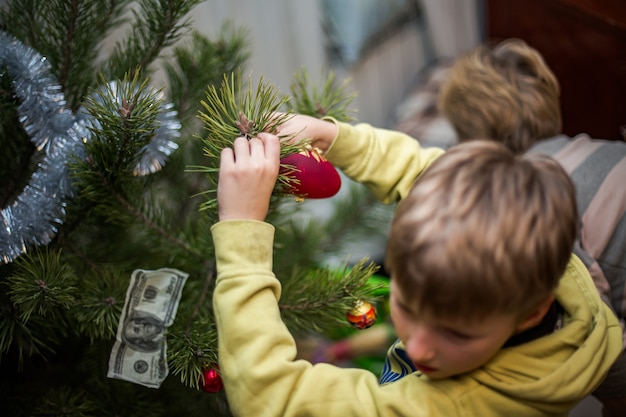 Jongen klampt zich thuis vast aan een rode bal aan een tak van een kerstboom. Thuis voorbereiden op Kerstmis
