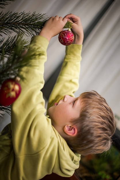 Jongen klampt zich thuis vast aan een rode bal aan een tak van een kerstboom. Thuis voorbereiden op Kerstmis