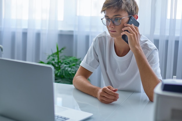 Jongen met bril praat aan de telefoon en kijkt naar een laptop