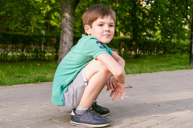 Jongen met donker haar gehurkt en kijkt bedachtzaam naar de kant. Kind hurkt op de stoep in het stadspark.