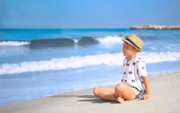 Jongen met hoed zit 's ochtends op het strand, op zoek naar golven