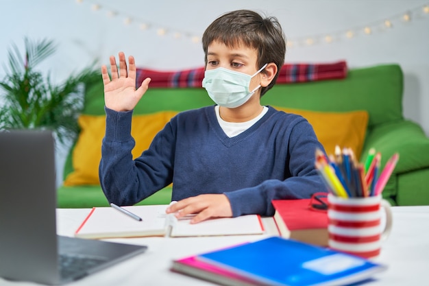 Jongen met masker met een videoconferentie op laptop met leraar vanuit huis, koptelefoon dragen en zwaaien op scherm, vrije ruimte