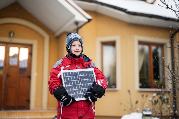 Jongen met zonnepaneel tegen huis in de winter Alternatief energieconcept