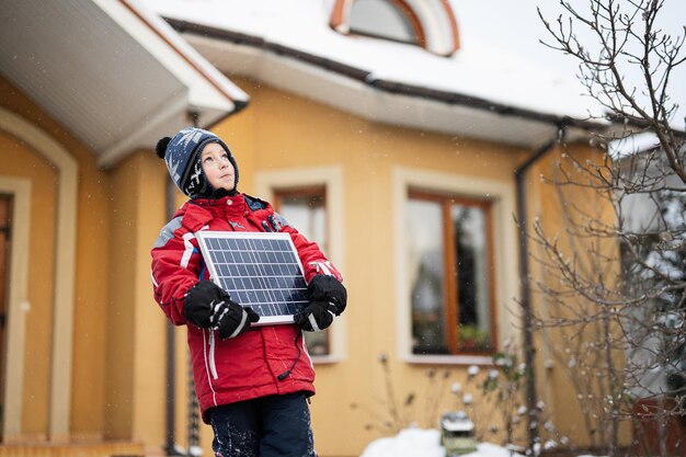 Jongen met zonnepaneel tegen huis in de winter alternatief energieconcept