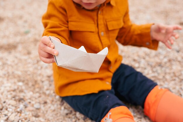 Jongen speelt met papieren boot met rubberen laarzen op het strand in de buurt van zee in de herfst of zomer