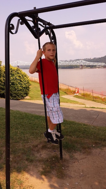 Foto jongen staat op buitenspeelapparatuur op een speeltuin bij de rivier