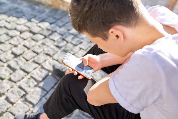 Jongen tiener 16,17 jaar oud, met behulp van smartphone, zittend op straat van zomer stad. Ruimte kopiëren, man leest een online bericht, bladert door sociale netwerken, typt tekst