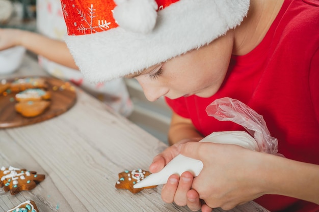 Jongen van 8 jaar oud in rode kerstmuts versiert peperkoekkoekjes met suikerglazuur op houten tafel in de keuken