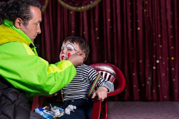 Jongen zit in stoel op leeg podium met clown make-up toegepast op gezicht door man, jongen kijkt naar camera uit hoek van oog