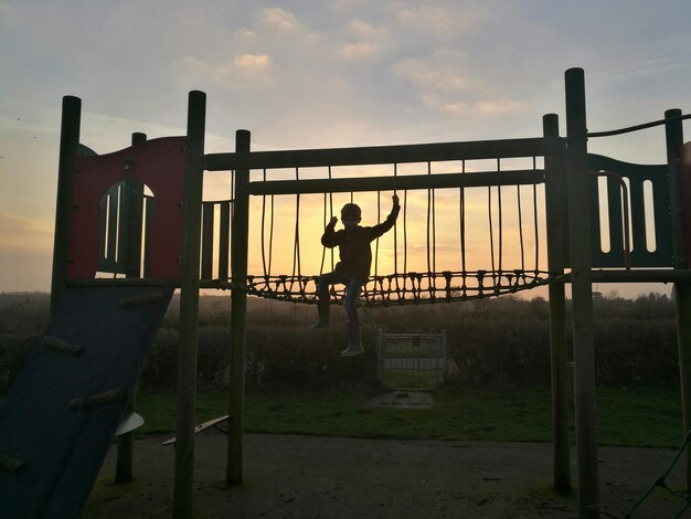 Foto jongen zit op buitenspeelapparatuur tegen de hemel tijdens de zonsondergang