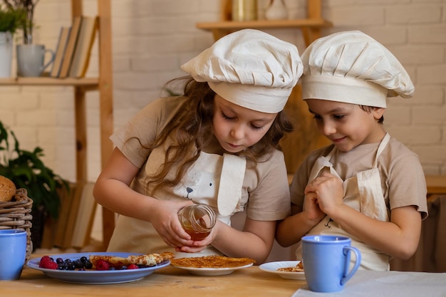 Jongens en een meisje aan tafel in schorten en koksmutsen gieten honing op pannenkoeken