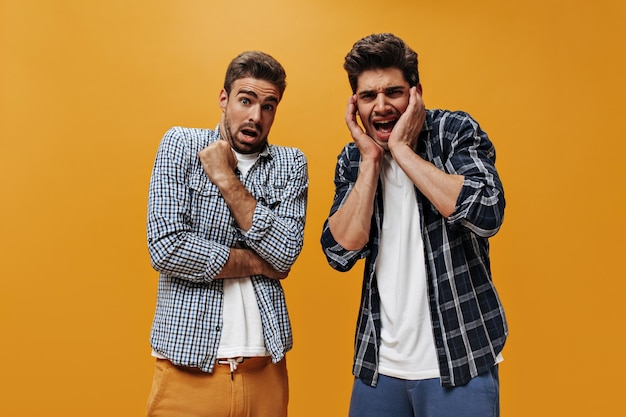 Jongens met baard voelen zich ongemakkelijk Emotionele brunette jonge vrienden in stijlvolle geruite shirts kijken in de camera op oranje achtergrond