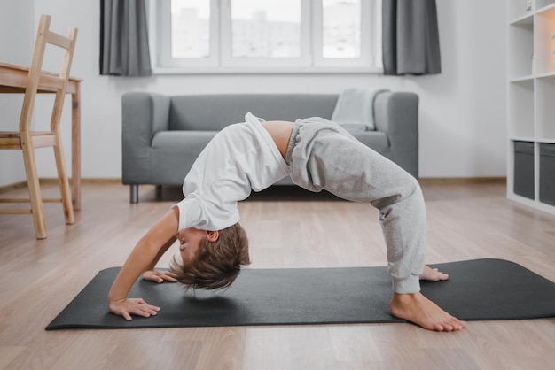 Jongenskind dat yoga beoefent, staande in brugoefening, urdhva dhanurasana pose, trainen, huiskleding, grijze broek, wit t-shirt, volledige lengte binnenshuis, interieur.