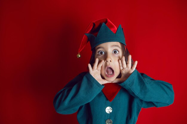 Jongenskind in een dwergkostuum staat op een rode achtergrond op eerste kerstdag