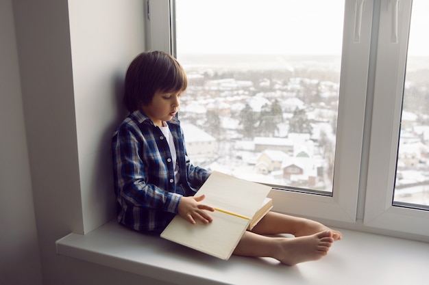 Jongenskind zit op het raam van een huis met een boek in de winter op een hoge verdieping