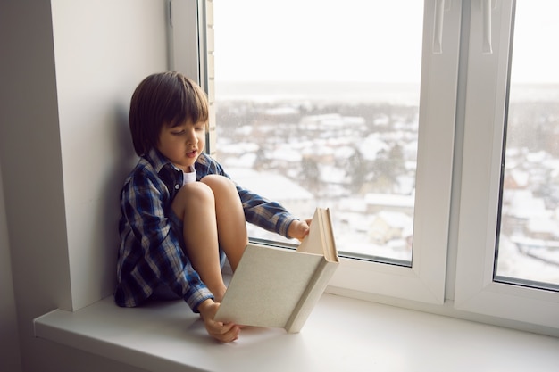Jongenskind zit op het raam van een huis met een boek in de winter op een hoge verdieping