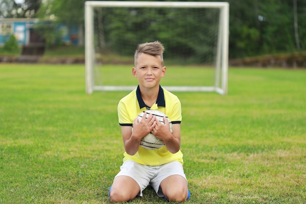 Jongensvoetballer zit op het voetbalveld tegen de achtergrond van het doel met de bal