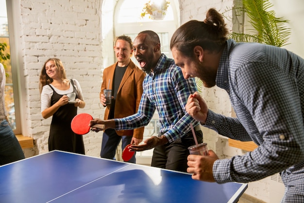 Jongeren die tafeltennis spelen op de werkplek, plezier hebben. Vrienden in vrijetijdskleding spelen samen pingpong op een zonnige dag. Concept van vrijetijdsbesteding, sport, vriendschap, teambuilding, teamwork.