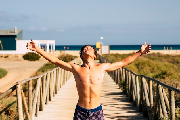 Jongeren en geluk in de zomervakantie met een knappe tiener die zijn armen opent en de frisse lucht inademt op het strand