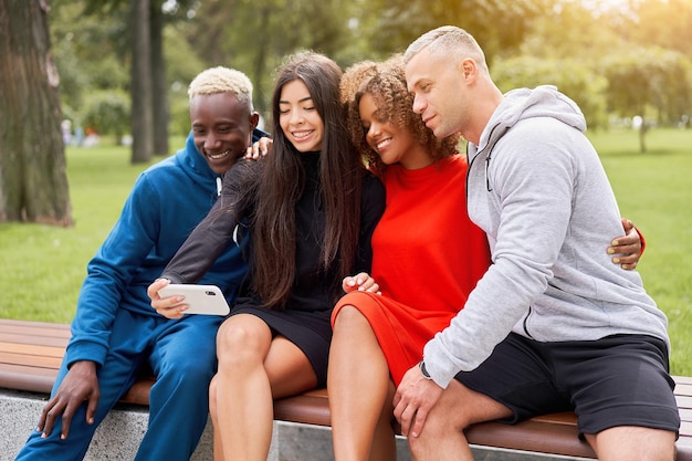 Jongeren van verschillende nationaliteiten nemen selfies met smartphones, zittend op een bankje in het park in de zomer. Atletische bouw. Sport uniform. Jonge gelukkige studenten.