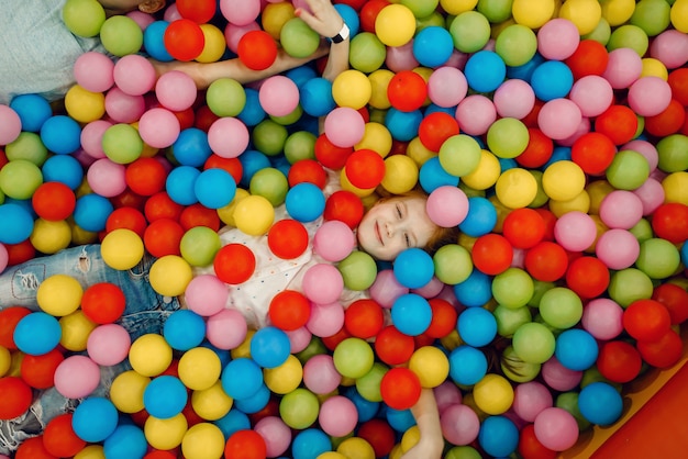 Jouful girl lying among many colorful balls in the entertainment center, top view. Female child leisures on holidays, childhood happiness, happy kids on playground