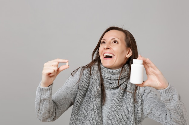 Jouful jonge vrouw in grijze trui, sjaal opzoeken, medicatie tabletten, aspirine pillen in fles geïsoleerd op grijze achtergrond te houden. Gezonde levensstijl ziek zieke ziekte behandeling koude seizoen concept.