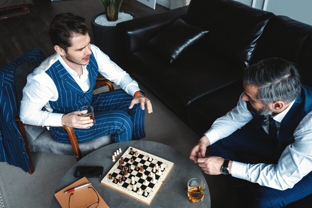 Jouw beurt! Twee jonge knappe mannen in volle pakken schaken en glimlachen terwijl ze binnenshuis zitten.