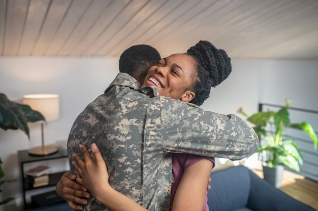 Joy of meeting. Cheerful dark skinned young woman with closed eyes hugging military man standing with back to camera at home