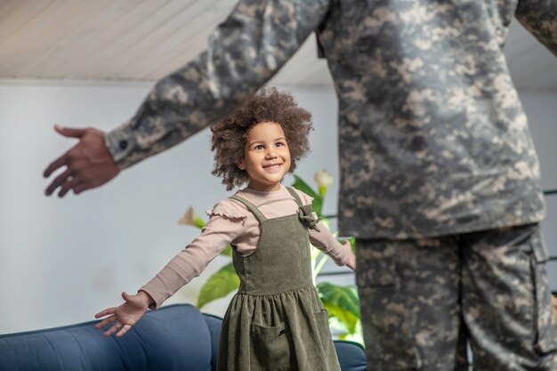 Joy, meeting. Dad in military uniform with back to camera and dark-skinned little happy daughter holding hands to sides during meeting