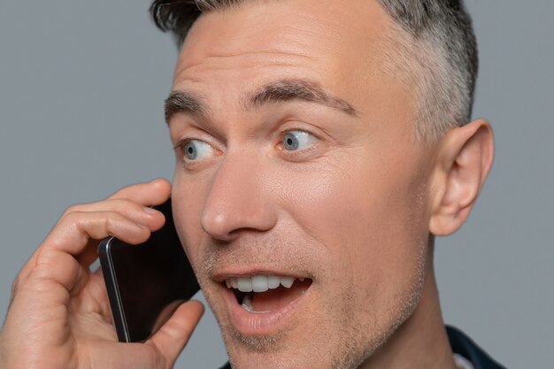 Joy, surprise. Close up face of joyful surprised man with gray eyes talking on smartphone