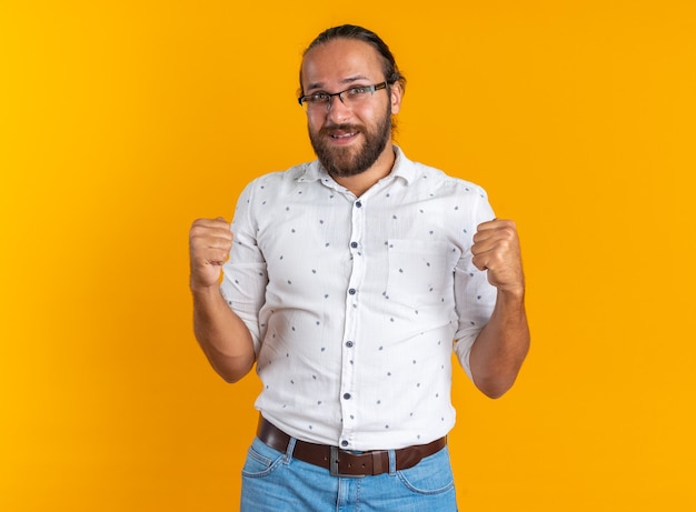 Joyful adult handsome man wearing glasses looking at camera doing yes gesture isolated on orange wall