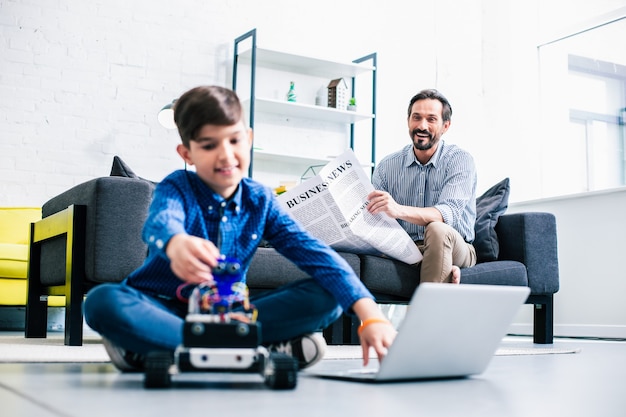 Joyful adult man reading a newspaper while his son testing robotic device