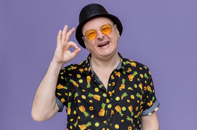 Joyful adult slavic man with black top hat wearing sunglasses gesturing ok sign 