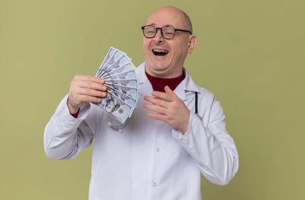 Joyful adult slavic man with optical glasses in doctor uniform with stethoscope holding money 