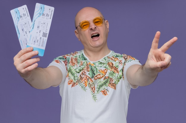 Joyful adult slavic man with sunglasses holding air tickets and gesturing victory sign