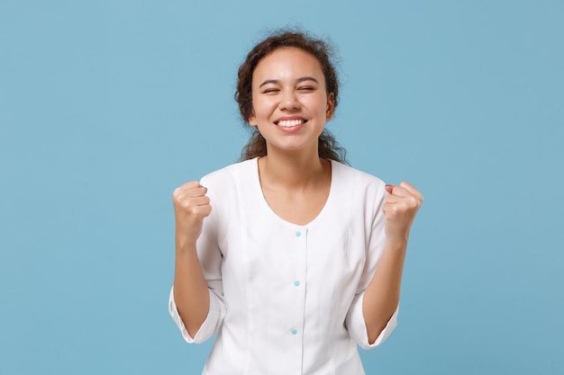 Joyful african american doctor woman isolated on blue\
background in studio. female doctor in white medical gown doing\
winner gesture. healthcare personnel health medicine concept. mock\
up copy space.