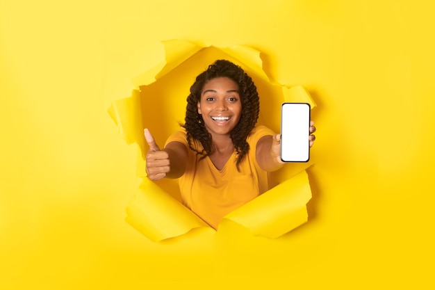 Joyful african american lady holding smartphone with blank screen and showing thumb up posing through torn hole in paper