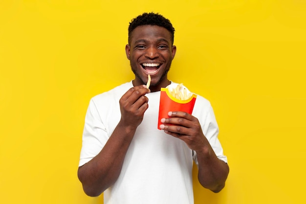 Joyful african american man holds french fries and bites snack on yellow isolated background