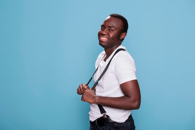 Uomo afroamericano gioioso che sorride cordialmente mentre posa per la macchina fotografica su sfondo blu. ragazzo dall'aspetto allegro e sicuro di sé che indossa abiti fashing mentre tira le bretelle.