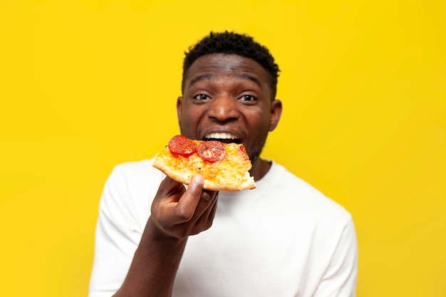 Photo joyful african american man in white tshirt bites piece of pizza on yellow isolated background