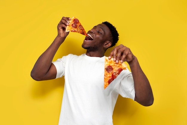 Joyful african american man in white tshirt eats piece of pizza and offers you on yellow background