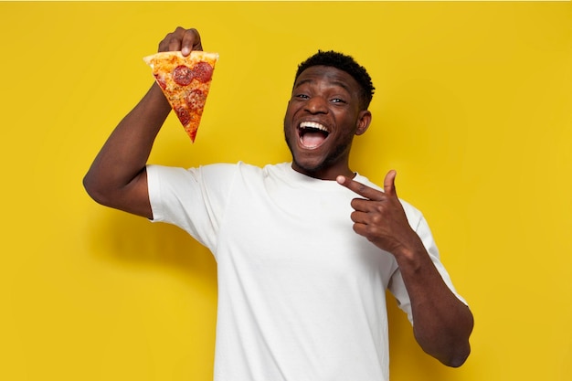 Joyful african american man in white tshirt holds piece of pizza and points his finger at it