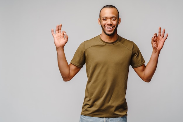 明るい灰色の壁にokのしぐさを示す緑のtシャツでうれしそうなアフリカ系アメリカ人の若い男