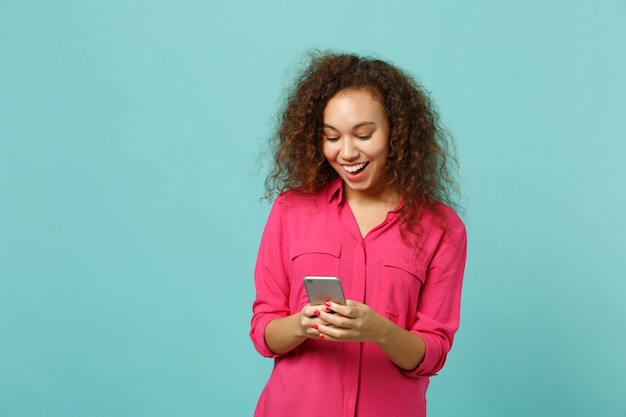 Joyful african girl in pink casual clothes using mobile phone, typing sms message isolated on blue turquoise wall background in studio. People sincere emotions, lifestyle concept. Mock up copy space.